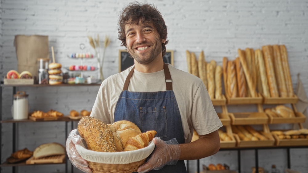 Hübscher junger Mann, der in einer Bäckerei lächelt, während er einen Korb mit frischem Brot hält, mit Regalen mit Backwaren im Hintergrund